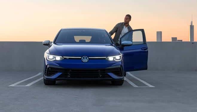 Man hopping into a blue Volkswagen vehicle at sunset in a city
