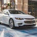 A white 2018 Chevy Malibu is driving in a city. The Malibu is a popular example of used cars near Atlanta GA being purchased.