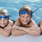 A row of children hanging onto the edge of the swimming pool with goggles on as they smile for a photo while in the water