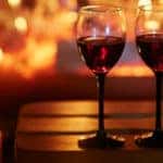 a couple of glasses of red wine set on a stool near lit candles and a fireplace