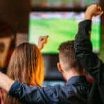 a group of friends huddle together at a sports bar with drinks as they cheer on their football team on the TV screen