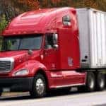 a red trailer and semi-truck driving on a country highway around a rocky cliff and next to an autumn forest