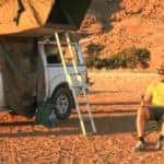 a couple sitting outside of a pickup camping truck with a ladder leading up to a tent on roof