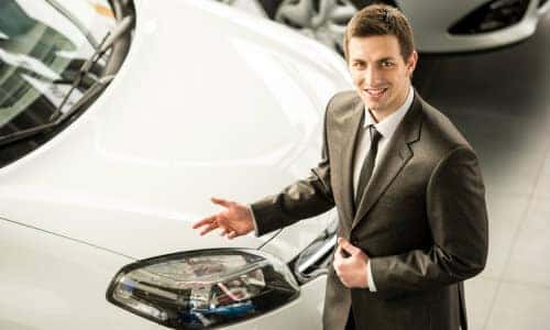 a car salesman standing next to a white car in a sales floor lot and looking up