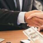 A handshake over a table with a dollar bill being passed