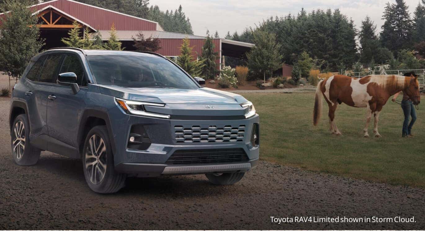 Front three-quarter view of a Storm Cloud Toyota RAV4 Limited parked on a dirt path next to a horse pasture and farm building.