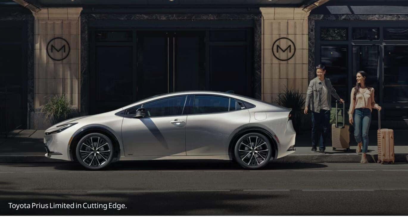 Left profile shot of a Cutting Edge Toyota Prius Limited parked curbside outside an upscale hotel as a couple with luggage approaches.