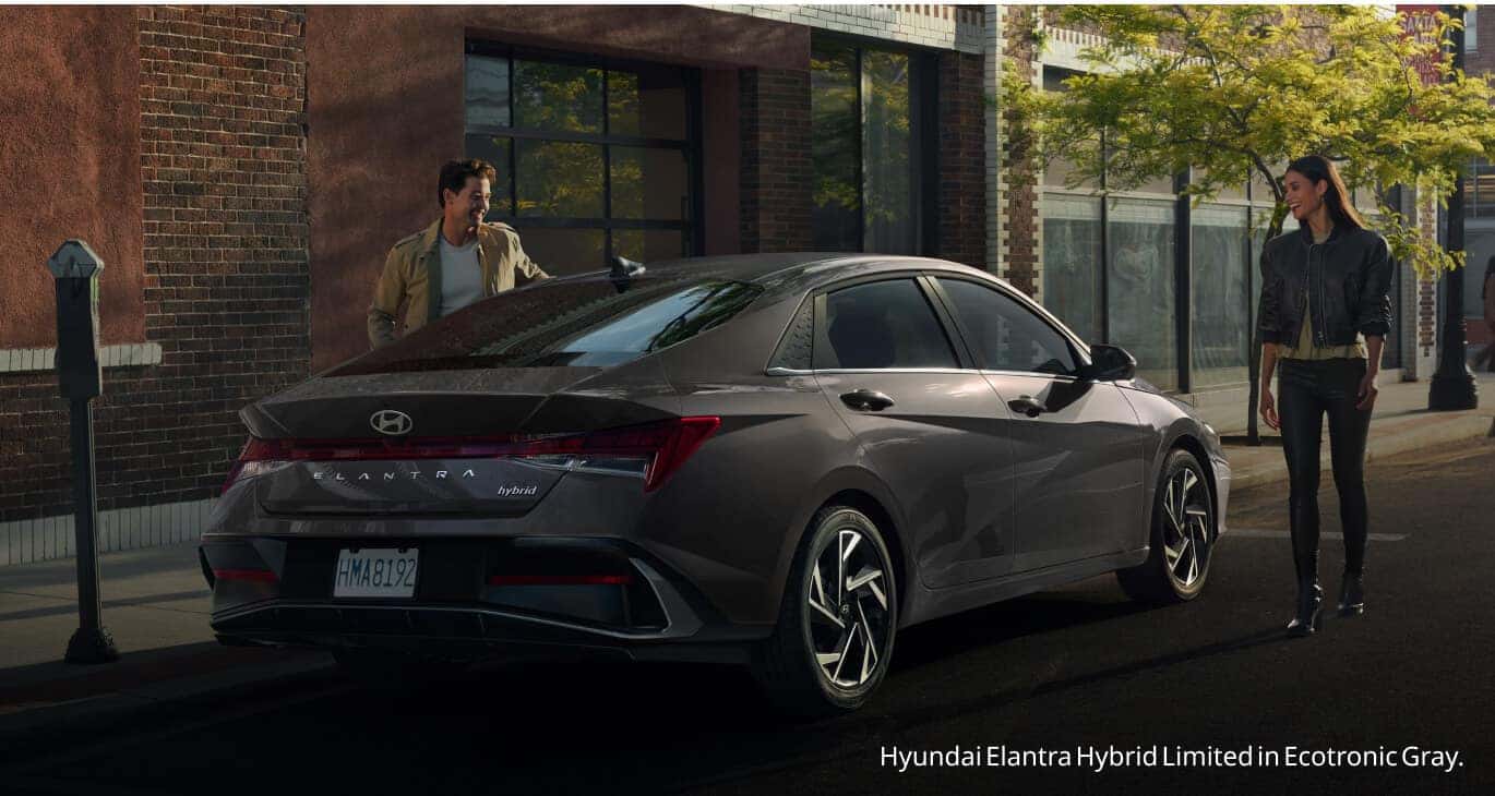 Rear three-quarter view of an Ecotronic Gray Hyundai Elantra Hybrid Limited parked curbside as a man opens the driver door.