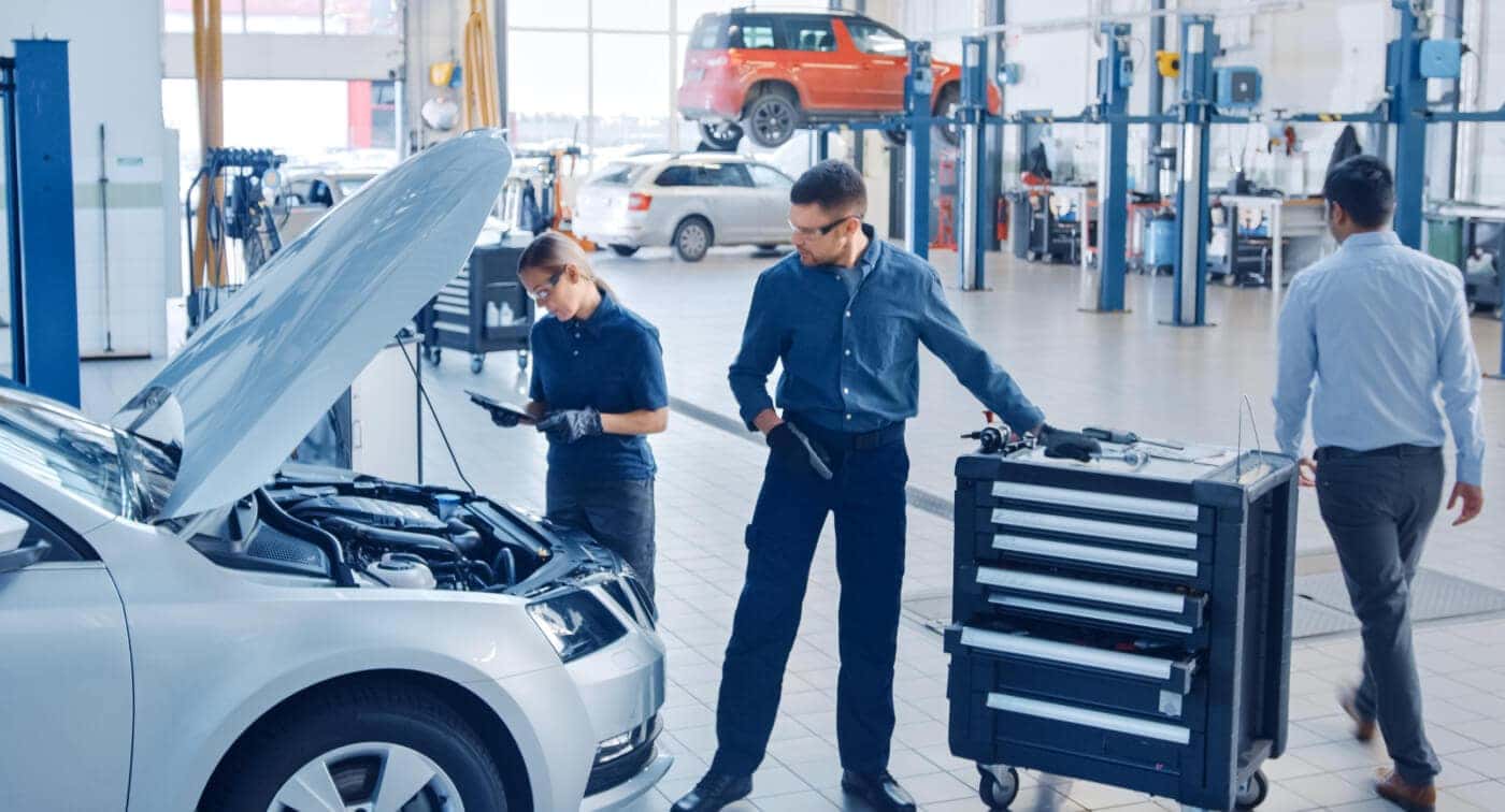 Wide angle view of three service technicians working under the hood of a car in a bright, clean shop.