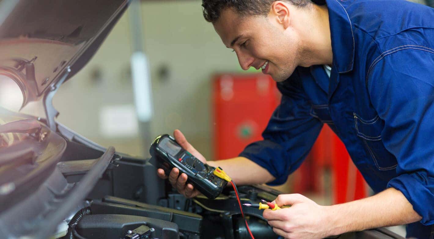 A man in a blue jumpsuit smiles while testing a car battery with a multimeter.