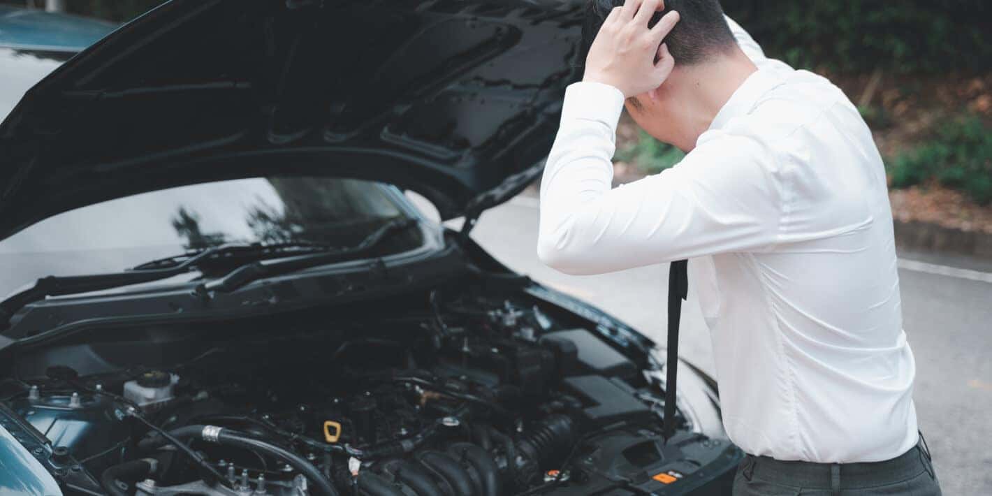 A man in a white shirt scratches his head while looking under the hood of a broken down car.