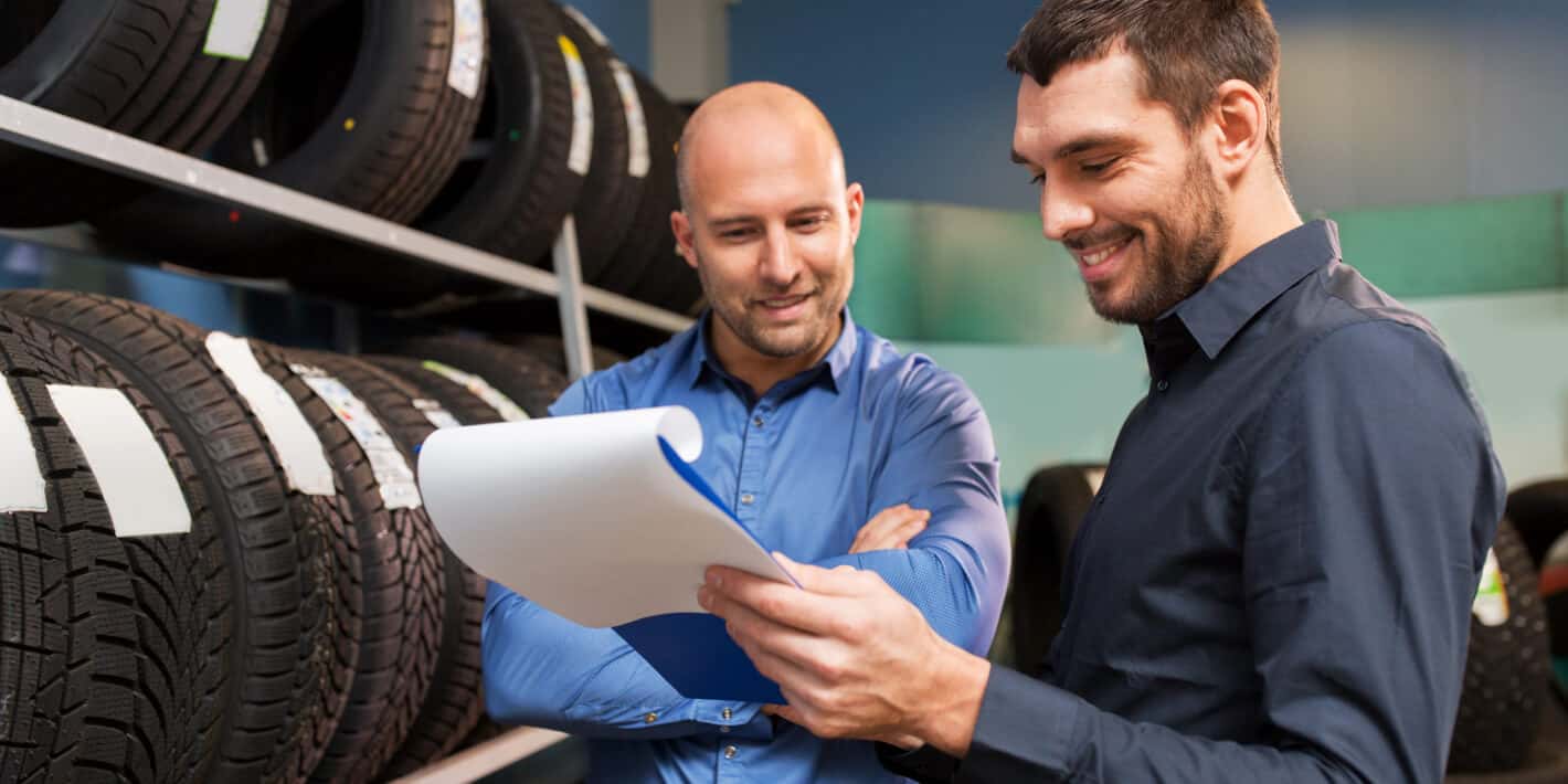 A service representative holding a clipboard assists a smiling customer in front of a rack full of tires.