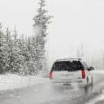 white SUV driving on tree lined highway with snow on ground and falling