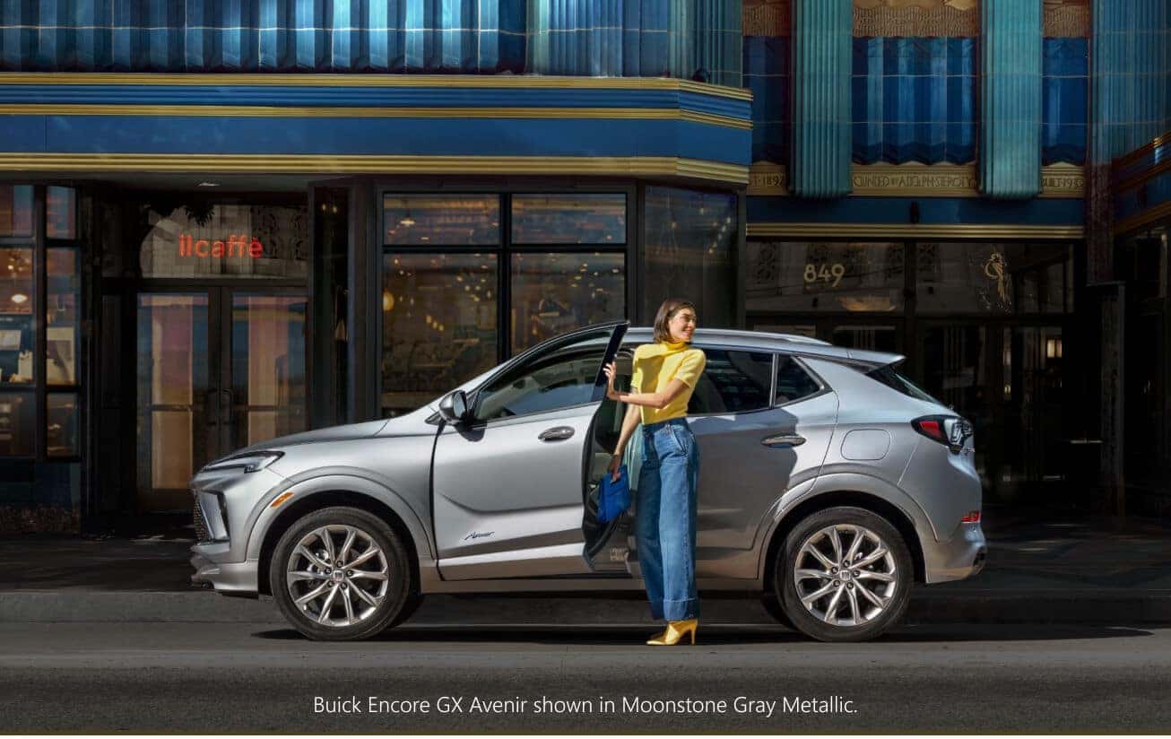 Side profile view of a silver Buick Encore GX Avenir being boarded by a woman in front of a  storefront.