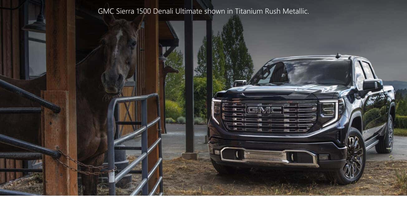 Front three-quarter view of a dark metallic GMC Sierra 1500 Denali Ultimate parked next to a horse stable.