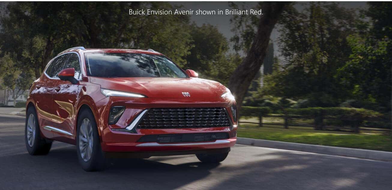 Front three-quarter view of a red Buick Envision Avenir driving down a tree-lined suburban street.