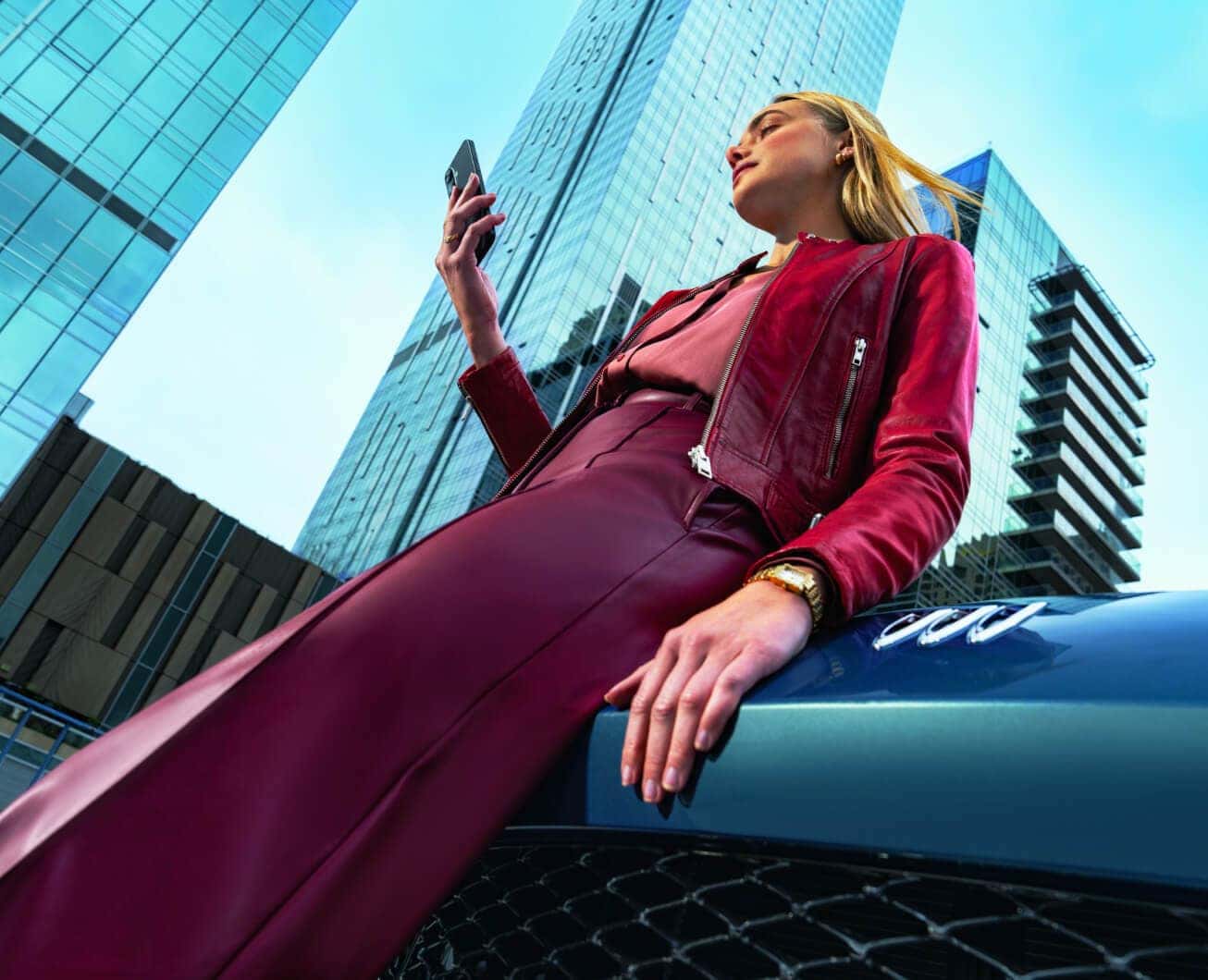 Low angle view of a woman holding a phone while sitting on the hood of a blue Buick Envision cityscape.