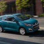 A teal 2019 Chevy Spark is driving in front of a brick building. It is part of the Chevy cars lineup.