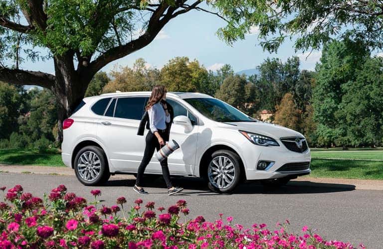 Woman walks alongside a white 2020 Buick Envision Essence parked under the bent branches of a tree.