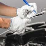 two white-gloved hands writing on a clipboard over a car engine
