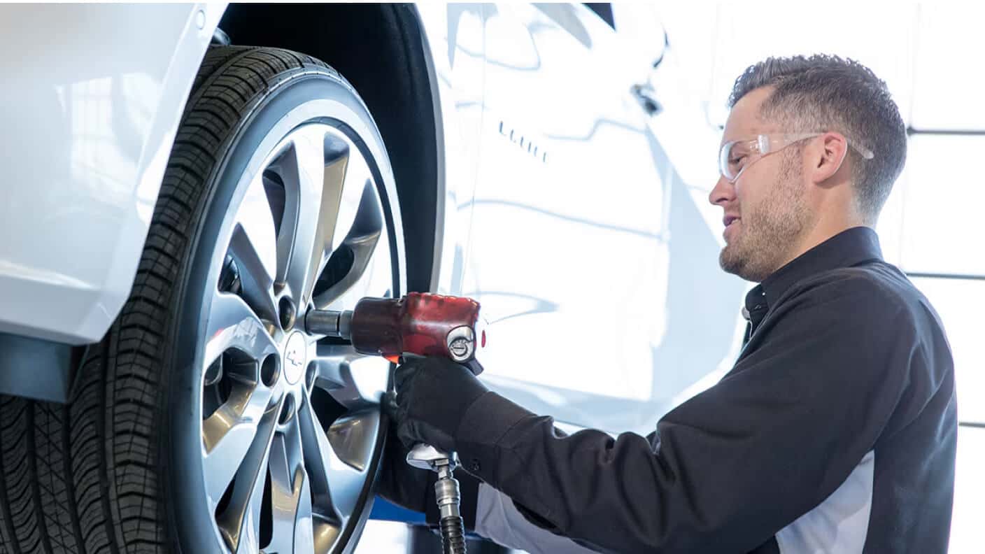 Close-up of a Chevrolet technician tightening the lug nuts of a Malibu with an impact wrench in a well-lit service bay.
