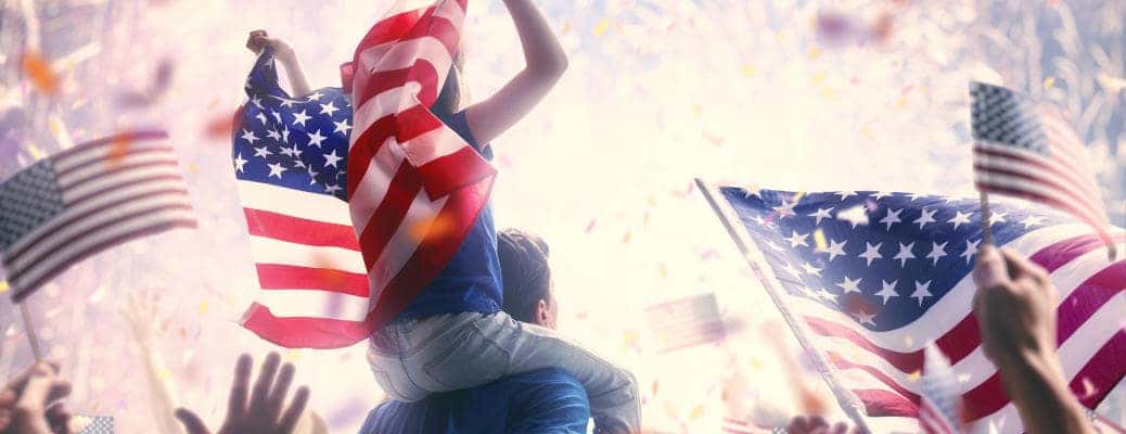 Patriotic holiday. Child sitting on shoulders of her father and holding the Flag of the USA. America celebrate 4th of July.