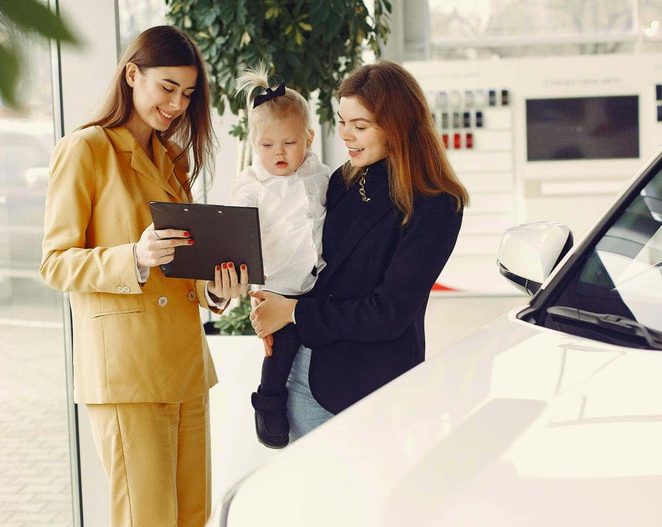 Dealership representative reviews used car options with a woman holding a child beside a white vehicle.