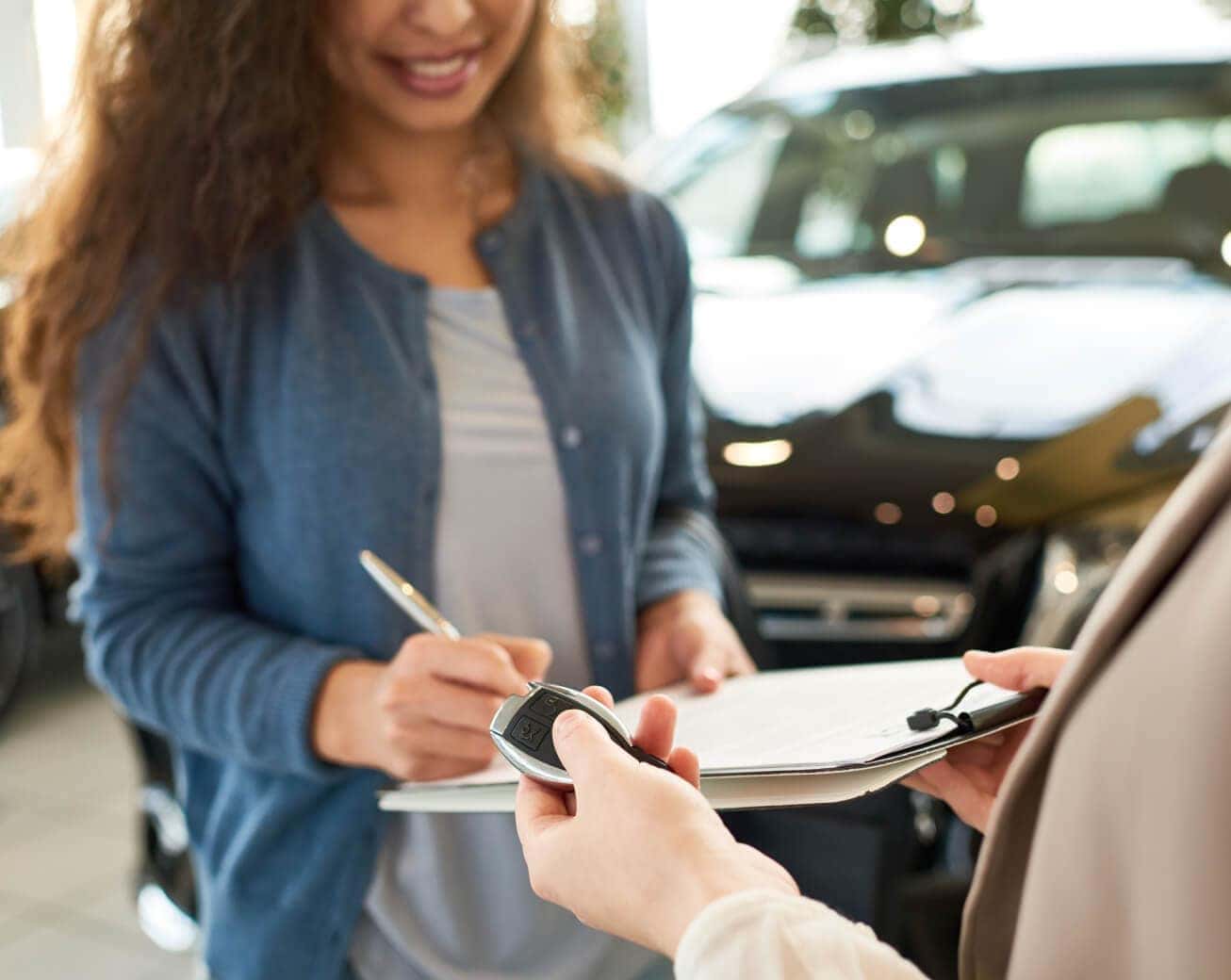 Customer signs financing paperwork as a dealership representative hands over a car key beside a vehicle.