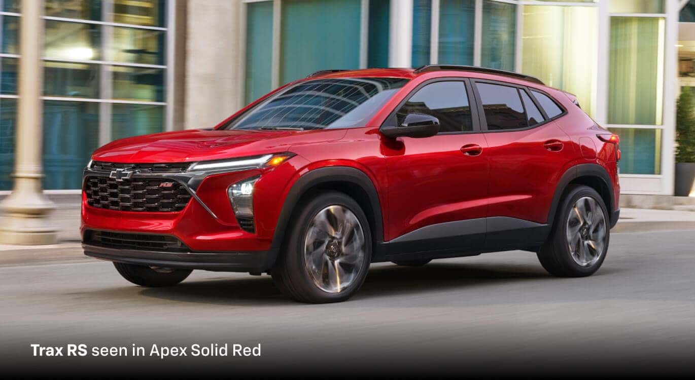 2026 Chevrolet Trax RS in Apex Solid Red, front three-quarter view driving past modern offices.