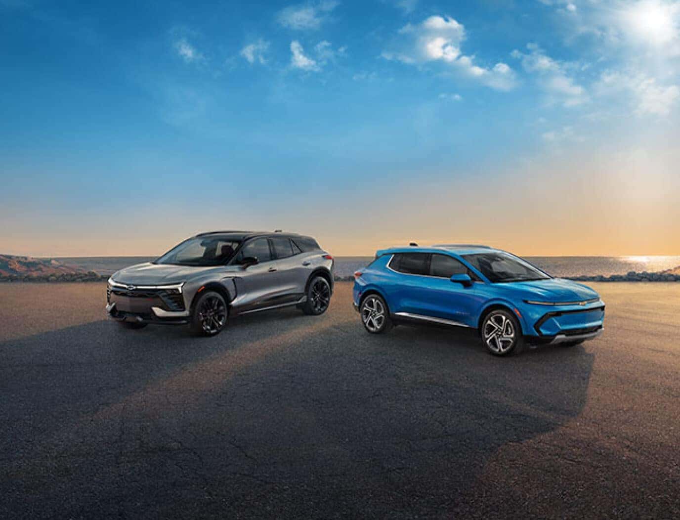 Front three-quarter view of silver Chevy Blazer EV and blue Equinox EV parked near ocean at sunset.