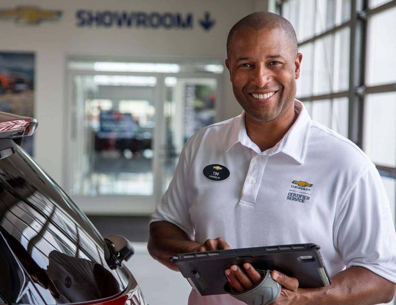 Portrait of a Chevrolet service advisor with a tablet next to a car inside a dealership showroom.