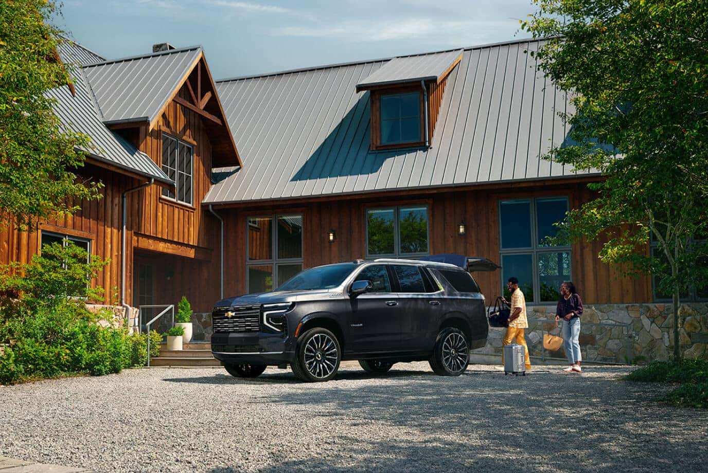 Front three-quarter view of dark gray Chevy Tahoe High Country parked at a wooden cabin driveway.