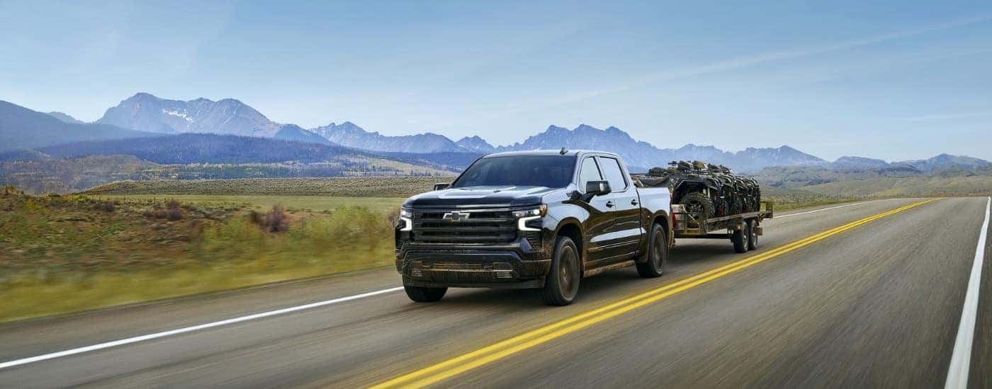 A black 2025 Chevy Silverado 1500 is shown from the front while towing ATVs.