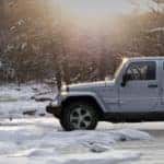 White Jeep Wrangler parked on icy terrain in a forest with the sun setting behind it