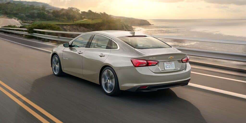 A Chevy Malibu drives down a coastal highway with the sun setting and light reflecting off the hood of the car.
