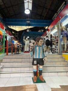 Inside a marketplace with a large caricature statue of a soccer player, with various stalls in the background.