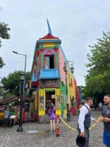 A street corner in Buenos Aires showing the colorful architecture of the La Boca neighborhood with a bright blue sky and a few people walking by.