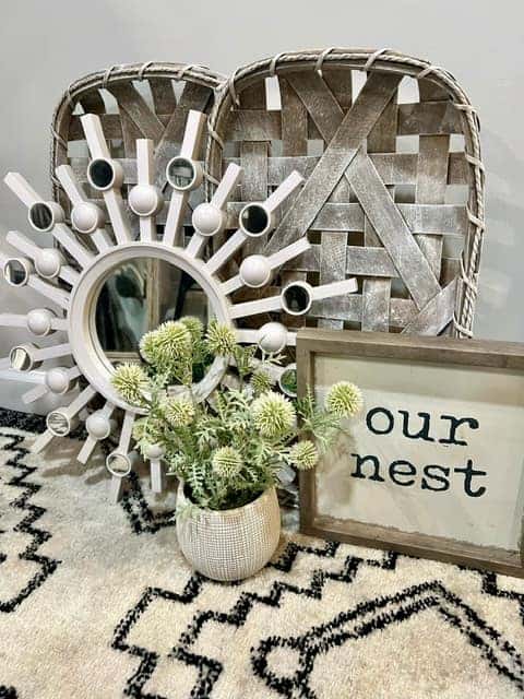A collection of home décor items including a decorative sunburst mirror, a framed sign saying "Our Nest," a small plant, and two woven baskets, donated to The Friends Program as part of the 100 Acts of Kindness initiative by Grappone Automotive Group.
