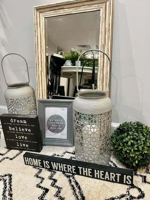 A collection of home décor items including two lanterns, a framed mirror, a small plant, and inspirational signs with messages like "Dream, Believe, Love, Live" and "Home is where the heart is," donated to The Friends Program as part of the 100 Acts of Kindness initiative by Grappone Automotive Group