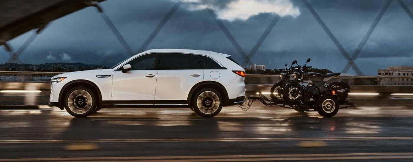 White 2024 Mazda CX-90 towing motorcycles across a bridge.