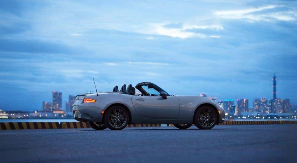 A light gray 2026 Mazda MX-5 Miata shown parked by water and a city.