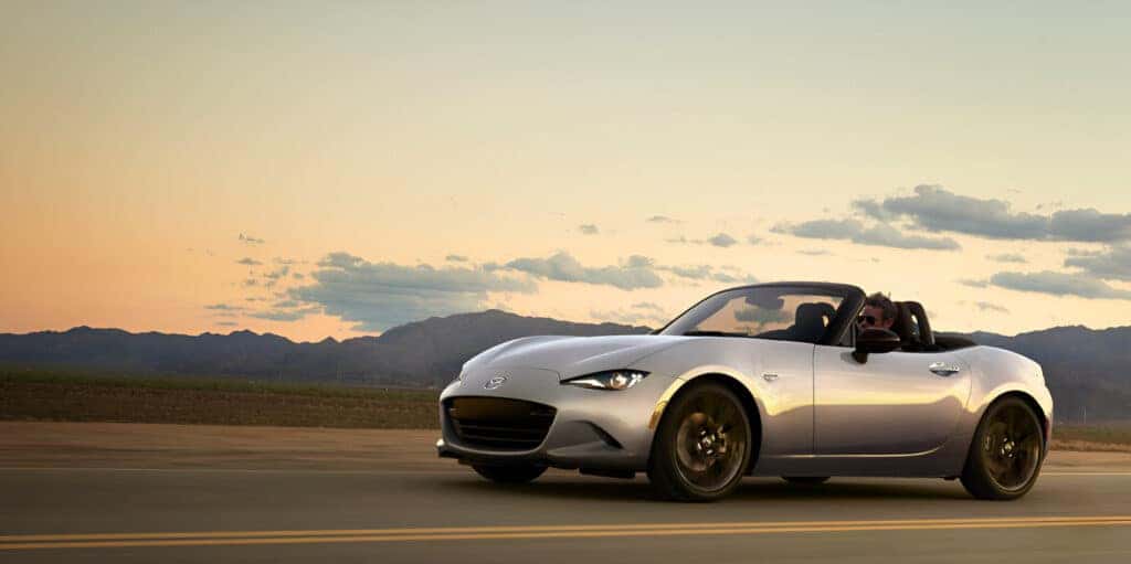 A 2024 Mazda MX-5 Roadster is cruising along a scenic road, showcasing its sleek design and sporty performance.