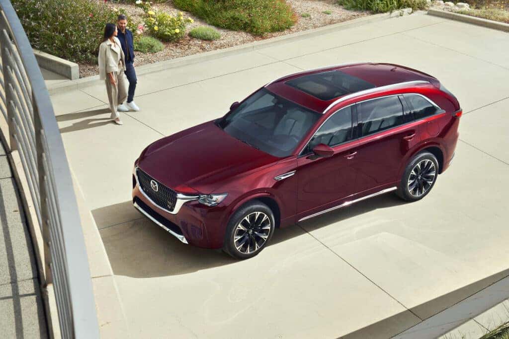 A 2025 Mazda CX-90 PHEV parked in front of a modern building, surrounded by lush greenery and rocky terrain.