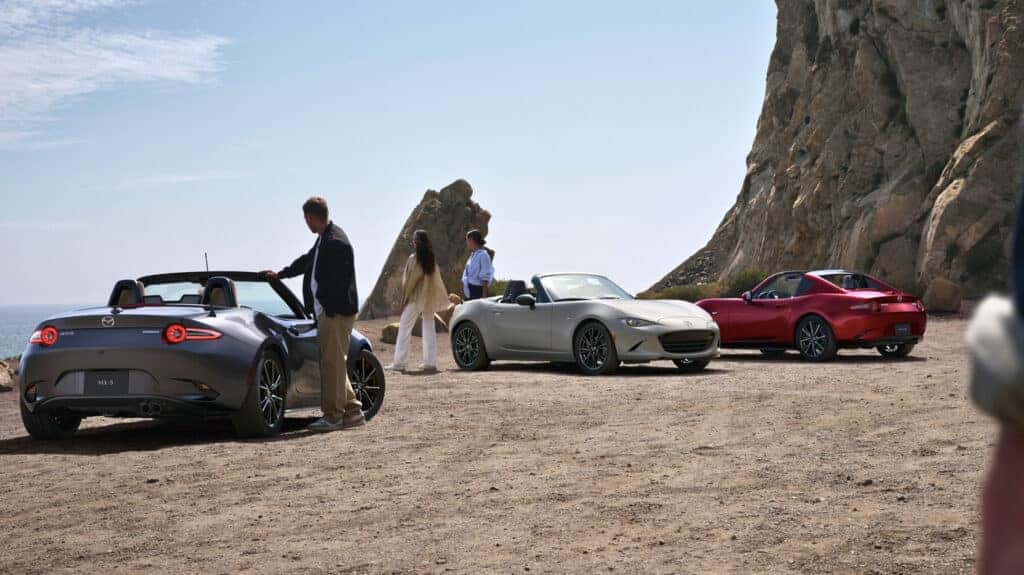 Three individuals pose beside their sleek Mazda Miata sports cars, showcasing their sleek and beautiful design.