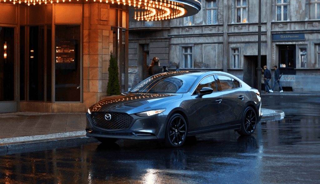 A 2025 Mazda3 sedan parked in front of a modern building, showcasing its sleek design against an urban backdrop.